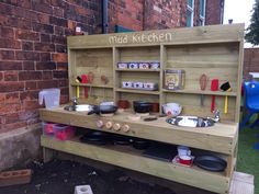 a wooden play kitchen with pots and pans on the shelves, in front of a brick wall