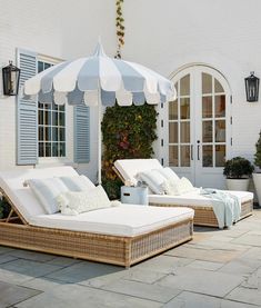 two chaise lounges with an umbrella in front of a white brick wall and blue shuttered windows