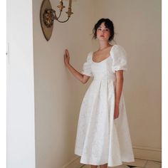 a woman in a white dress standing next to a wall with a clock on it