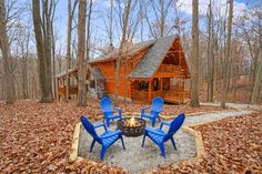 a fire pit surrounded by blue chairs in front of a log cabin with trees and leaves on the ground