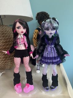 two dolls standing next to each other on a table with a lamp in the background