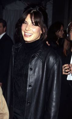 a woman wearing a black leather jacket and smiling at the camera with other people in the background