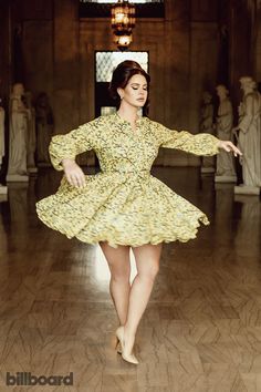 a woman in a yellow dress dancing on a wooden floor with statues behind her at the back