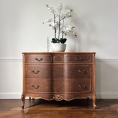 a vase with flowers on top of a wooden dresser next to a white wall and hardwood floor