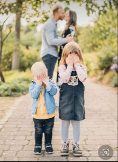 two children covering their eyes as they stand on a brick path in front of trees