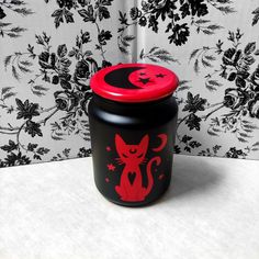 a black and red canister with a cat design on the lid sitting in front of a floral wallpaper