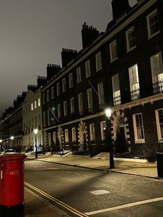 a city street at night with buildings on both sides and a red post box in the middle