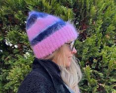 a woman wearing a pink and blue knitted hat with stripes on the side, standing in front of some trees