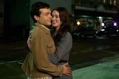 a young man and woman embracing on the street at night with cars in the background