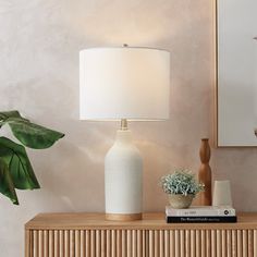 a white vase sitting on top of a wooden table next to a lamp and potted plant