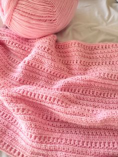 a pink crocheted blanket next to a ball of yarn on top of a bed