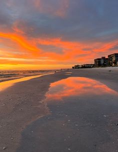 the sun is setting at the beach with buildings in the background and water reflecting on the sand