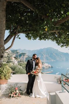 a bride and groom standing under a tree on top of a hill overlooking the ocean