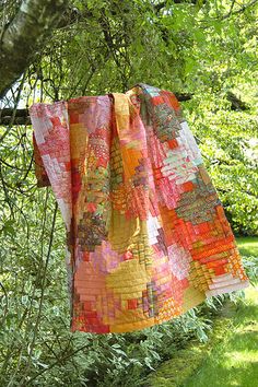 a patchwork quilt hanging from a tree in the shade by some grass and trees