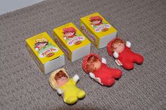 three little dolls sitting on the floor next to two boxes of cereal and one doll