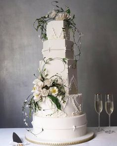 a tall white wedding cake with flowers and pearls on the top, surrounded by wine glasses