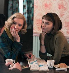 two women sitting at a table with coffee and pastries in front of them, talking to each other