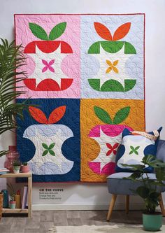 an apple quilt hanging on the wall next to a blue chair and potted plant