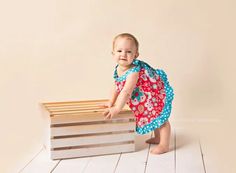 a baby girl in a red and blue dress standing next to a stack of wooden crates