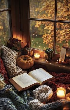 an open book sitting on top of a blanket next to candles and pumpkins in front of a window