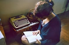 a woman sitting on the floor reading a book next to an old fashioned typewriter