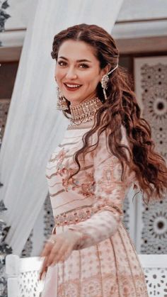 a woman standing in front of a white curtain wearing a pink dress and gold jewelry