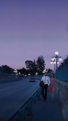 a man and woman are walking down the sidewalk at night with street lights in the background