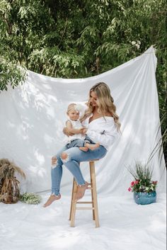 a woman holding a baby sitting on top of a wooden stool in front of a white backdrop