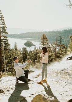 a man kneeling down next to a woman on top of a mountain near a lake