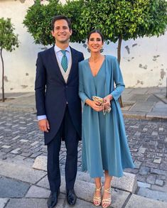 a man and woman standing next to each other in front of a tree on a cobblestone street