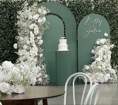 two white chairs sitting next to each other near a table with flowers on it and greenery behind them