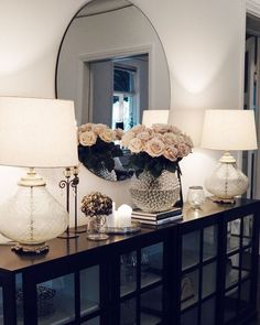 a table with two lamps and flowers on it in front of a large round mirror