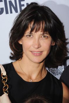 a woman with shoulder length hair holding a purse in her right hand and smiling at the camera