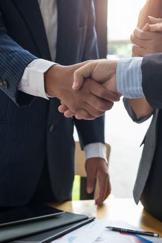 two men in suits shaking hands over a desk with papers and pens on the table