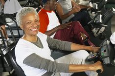 an older woman is smiling as she rides on a stationary exercise bike with other people in the background