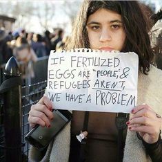 a woman holding up a sign that says if fertiized eggs are people & refugees aren't we have a problem