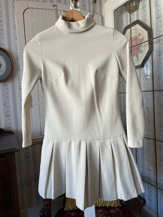 This unique dress is made from medium weight knit in classic beige. It has a high mock neck and a dropped waist with a pleated skirt. It fastens with a long zipper in the back. The measurements, taken with the dress lying flat, are: shoulder to shoulder, 14 inches; armpit to armpit, 16 inches; sleeves, 23 inches; waist, 15 inches; length, 34 inches; bottom edge,34  inches. In very good condition. Fitted White Mini Dress With Pleated Waist, Elegant Turtleneck Mini Dress For Spring, Fitted Beige Mini Dress For Fall, Fitted Beige Mini Dress For Winter, Beige Fitted Mini Dress For Fall, Beige Fitted Mini Dress For Winter, Winter Beige Fitted Mini Dress, Classic A-line Mini Dress For Fall, Chic High Neck Cream Dress