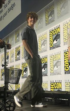 the young man is walking around in front of comic convention panel wall with his foot on a cart