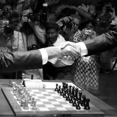 two men shaking hands over a chess board with people in the background taking pictures behind them