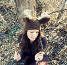 a woman sitting on the ground in front of a tree wearing a knitted deer hat