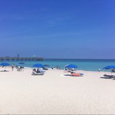 many people are on the beach with blue umbrellas and lounge chairs in the sand