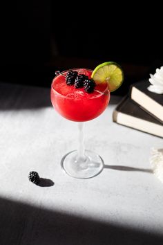 a red cocktail with blackberries and lime garnish sits on a table next to an open book