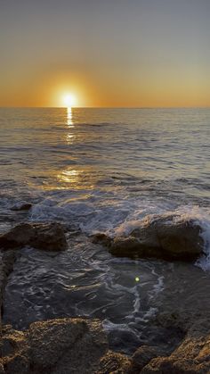 the sun is setting over the ocean with waves crashing on rocks in front of it