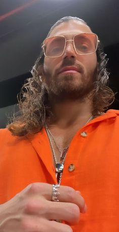 a man with long hair wearing an orange shirt and silver chain around his neck is staring at the camera