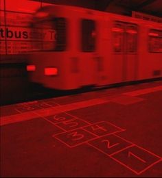 a train traveling down tracks next to a red lit subway station platform with numbers painted on the floor