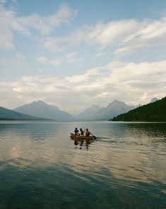 three people in a small boat on a lake with mountains in the background and clouds in the sky