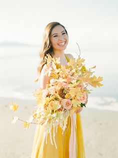 a woman in a yellow dress holding a bouquet of flowers on the beach with water in the background