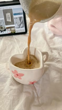 coffee being poured into a white cup on top of a bed next to a laptop