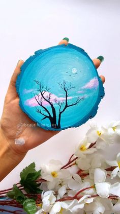 a person holding up a piece of wood with a tree painted on it and flowers in the foreground