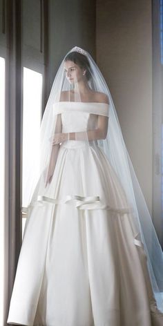 a woman in a white wedding dress standing by a window with her veil pulled back